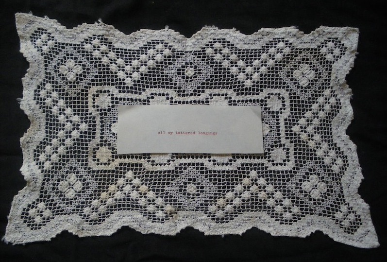 A cream vintage rectangle doily lays on a black background. Upon the middle of the textile piece is a cream piece of paper. Centered on the paper, in red typewritten ink, are the words 'all my tattered longings'.