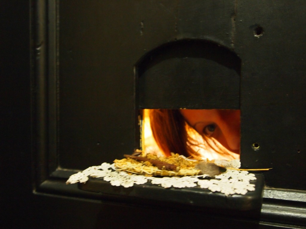 there is a small arched opening in a big black door. this is a close up shot of the opening, it has a little shelf in front of it with some old cream textile and little nature bits on it ... the opening is open, a poets head peeks out from the other side, we can see an eye a nose and some hair, warmly lit by the light behind.