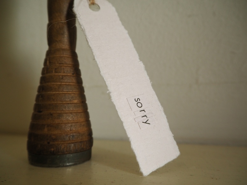 apology . detail .. the base of the vase sits on a creamy yellow surface with white wall behind ... its rich colour is contrasted by a creamy white tag hanging down from the vase's side to the right .. this side of the tag is made from a page reclaimed from the book .. it is blank but for one word which has been collaged together from letters carefully cut from the book’s print .. the word rest near the base of the tag ... the letters: so.r.r.y