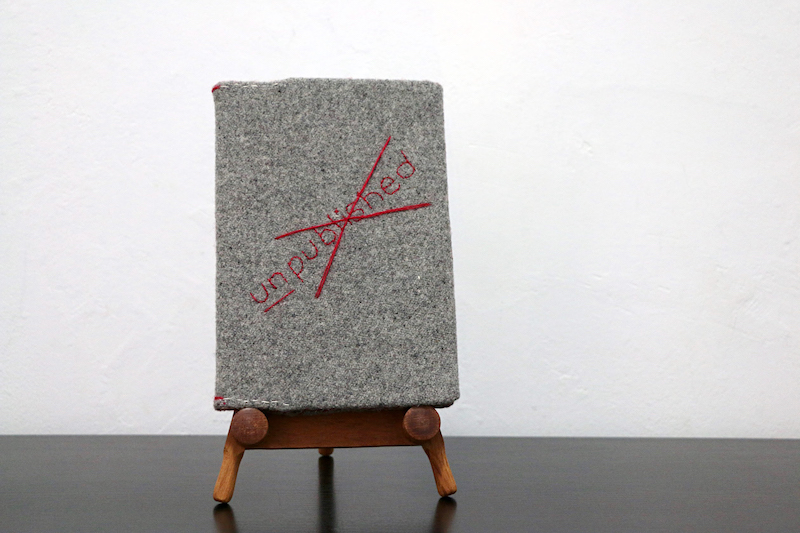 dust jacket artwork .. a full image of the work .. a book sits on a wooden book stand on a black surface in front of a white textured wall ... the wooden stand has a mid-range stain, slightly rounded legs and the book rests upon two wooden circles affixed to its front ... the book has been covered with grey woollen fabric ... on the front of this handstitched dust jacket, the word unpublished is stitched in red thread .. the ‘un’ is underlined .. the ‘published’ is covered by a horizontal red stitched cross