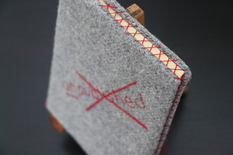 dust jacket artwork from above .. a grey wool-covered book, against a black background ... on the front a red word is blurred by the camera .. the word is covered by a red cross ... all along the top and side of the book the open edge is crisscrossed by repeated red thread crosses .. stitches holding the front and back of the protective cover closed .. the paper edge is visible behind the red threads
