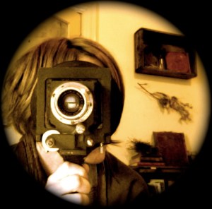 this photo is circular. in a glowing circle with a black edge ... indigo is holding a vintage camara lens structure to her eye ... it covers almost her whole face, she has short dark blonde hair cut in layers, rounding her head ... the lens is round glass reflecting two offset rectangles of light, surrounded by black circle with tiny digits or markings, by silver circle, by black square surround, with silver knobs and adjusters ... held in a comfortable grip by indigo's hand on a vertical handle or some such, from the base. in the soft glowing background there is a frosted glass door, slightly open, wooden box holding fossicked treasures on the wall, below that directly on the wall is a found twig with birds nest, and under that a wooden army crate turned book shelf with books and nests and other collected items within and on top. photo: indigo eli