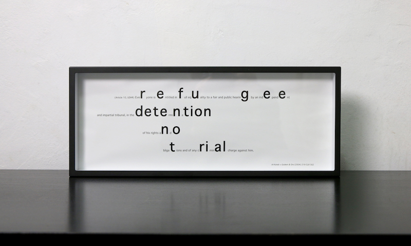 in.definite artwork .. a full image of the work ... a thin rectangular horizontal black frame is itself framed by the black surface it is resting on and the white textured wall behind .. inside the shadow box frame is black text on a white background .. some of the text is reflected in the black surface below ... inside the frame, Article 10 of the Universal Declaration of Human Rights is printed .. there are several off-centred lines of text .. some letters are enlarged to form new words from within the text .. the enlarged letters read: refugee detention no trial .. in the bottom right of the frame is a strip of text, a citation of a legal case: Al-Kateb v Godwin & Ors (2004)