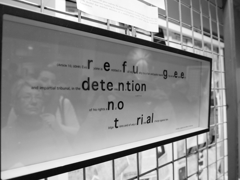in.definite artwork on display .. a full image of the work viewed from the side, a horizontal rectangular dark shadow box frame with text inside, black on white ... the frame is hanging on a wire mesh wall ... the rounded font reads (Article 10, UDHR) everyone is entitled in full equality to a fair and public .. the words trail off to the right .. within the printed article letters have been enlarged and form another message: refugee detention no trial .. reflected in the frame’s glass are the faces of several people audiencing the work .. a woman with short hair and glasses looks up to consider the work .. a younger woman with long hair stands behind her, an arm upon on her shoulder