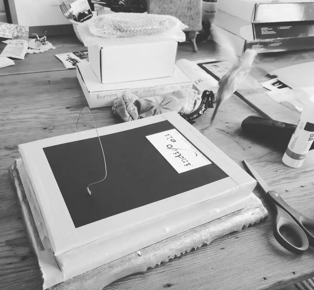 black and white photo of indigo's work table ... a framed artwork is face down being taped and secured back up after an update ... the table is covered with art pieces and boxes of artworks, scissors, a stick of glue ... from the table, a budgie is launching off into flight