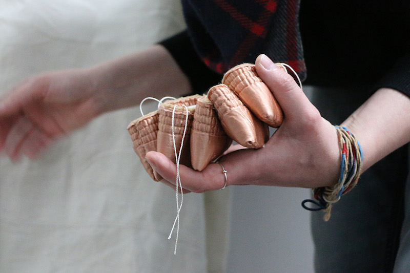 luxury artwork .. a close up shot of indigo holding 5 pieces within the soft stretch of her left hand ... she is wearing a black jumper pulled up at the forearms ... on her index finger is a thin silver ring with a silver bauble .. a wrapped threaded bracelet adorns her left wrist with hues of blue white red and gold .. a tartan scarf . dark blue and grey with red highlights . hangs into the shot from her neck ... her right arm is blurred from movement, reaching toward something out of shot ... the 5 works in her hand are small satin purses in ballet slipper pink, made from woollen blanket edging .. draping from some of the purses are doubled white threads