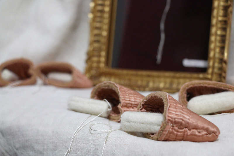 luxury artwork full shot ... a textured gold edged shadow box frame sits on cream-white linen .. pink satin purses are laid out in front, woollen tampons with satin white strings are tucked in or spilling out ... in the frame, is one cream-white cashmere tampon, silky strings draping down the deep red velvet background to the bottom of the frame where a white paper label is attached in the right corner .. the text on the label is unclear