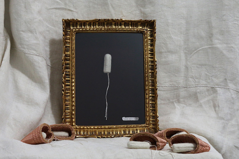 luxury artwork full shot ... pinky blanket-edge purses are laid on a cream-white linen fabric in front of a a textured gold edged shadow box frame .. in front of the frame 5 pinkish satin purses lay open .. their contents tucked in or spilling out .. fluffy cream-white woollen tampons with shimmering white strings ... in the frame presented on a deep red background is one cream-white woollen hand felted tampon, silky strings draping to the bottom of the frame where a white paper label is attached in the the right corner .. the text is are unclear