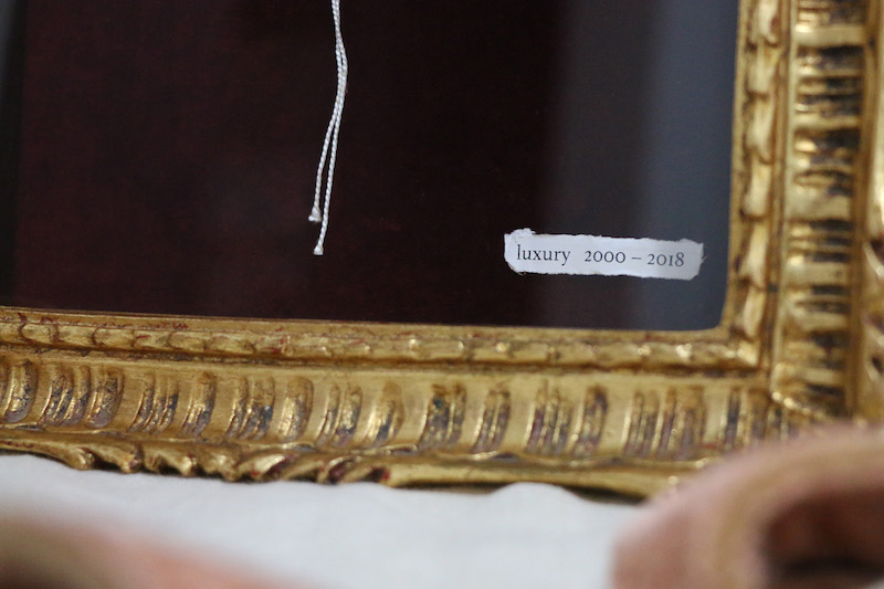 luxury artwork detail ... a close up of the bottom edge of a textured gilt frame with pink satin purse edges in front, blurred by the camera’s depth of field ... inside the frame, in the middle of a dark background, hang two silky white threads .. in the bottom right corner of the frame a torn paper label has been stitched in, it reads : luxury 2000 – 2018