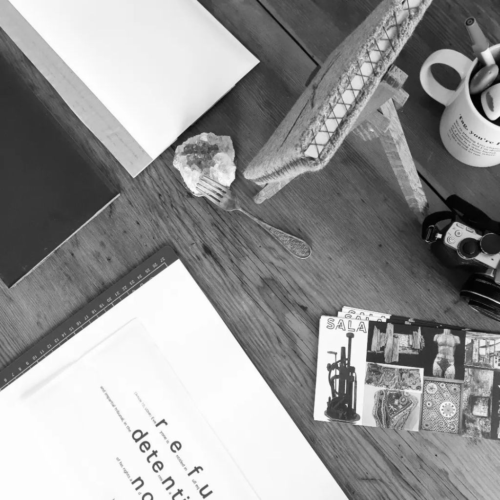 Photo by indigo eli on July 08, 2023. May be a black-and-white image of diary, clipboard, pen, book, typewriter, desk and text. Or may be that that was a robot interpretation of an artist's worktable with an deconstructed artwork, a stitched something, a poetry mug full of highlighters, a camera, some SALA flyers, and an alphabet fork resting on a hunk of amethyst crystal.