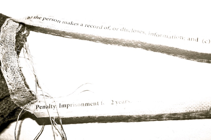 blanket silence artwork detail .. a black and white image of the work .. a section showing the gauze ties stretching out from the right side of the mask ... the gauze ties .. made from one long piece .. are attached along the side of the grey wool mask with hand stitches that blend in with the white fabric mesh over the dark grey .. the ties pulling away from top and bottom of the mask to the right .. have black text attached .. this close up reveals that the words are printed on transparent paper that has been sewn on to the ties .. the words blur off to the right .. the shadow of the ties is pronounced and textural underneath ... the top tie reads: (b) the person makes a record of, or discloses, information; and (c) .. the bottom: Penalty Imprisonment .. 2 years.