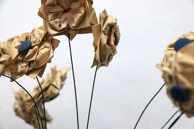 apology .. a close up detail of folded paper flowers fanned out on thin, dark wire stems in front of a white background ... on the sides of the image, groups of flowers are blurred by the cameras focus .. near the centre, 3 flowers stand tall and clear ... folded from aged yellow book pages, these flowers are small and dense, layered, 3 dimensional .. their petals decrease in size towards the middle .. one has a dark blue fabric button at its centre .. another . the tallest . is centred by stitches of woollen red thread ... snippets of text are revealed on the folded petals, folded pages ... words like: ‘wished’ .. ‘up’ .. ‘true’