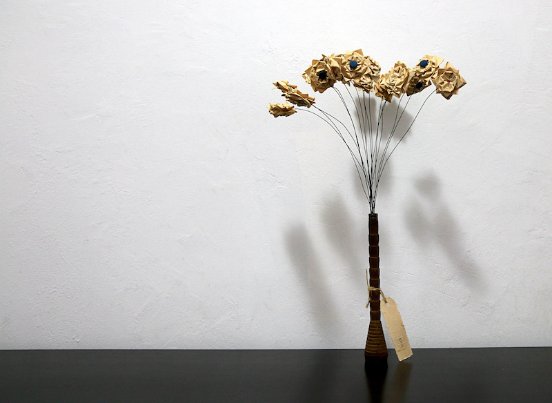 apology .. a full image of the work .. a thin vase of flowers sits to the right of frame on a black surface in front of a white textured wall ... the wooden vase, a repurposed industrial bobbin, has a short conical base that straightens into a tall thin cylinder with notches up its spine .. a tag, thin and rectangular, with a hole punched in one end, is tied to the vase by twine, on it, some indistinguishable text ... at the top of the vase, long thin wire stems fan out to hold a bunch of paper flowers folded from book pages. From right to left their faces point out toward the viewer . point up . tilt to one side, to the other . face the viewer ... the three on the very left tilt face down on slightly curved stems ... some flowers have round blue fabric buttons at their centre .. some have a hint of red thread instead ... on the wall behind are shadows of the flowers .. organic blobs form a loose arrow shape near the base of the work .. shifting into star-like shapes at the top