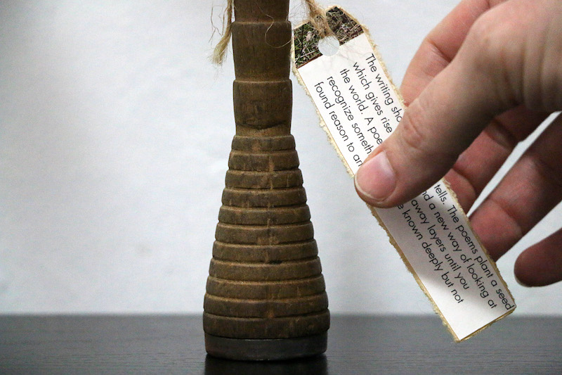 apology artwork, detail of the base of the vase . a wooden industrial bobbin from the old Onkaparinga woollen mill . is sat on a black surface with white wall behind ... its conical base is ribbed by evenly spaced carved lines before it straightens into a thinner spindle .. with notches spaced further apart ... its rich colour is both stained and lightened, by age and wear … hanging from the top of the image are two ends of natural twine .. one is looped through a hole in a long rectangular tag ... this side of the tag is shiny, a clipping from the back cover of a book containing the artist’s unlicensed work .. it is edged by softly torn edges from the paper layer behind ... at the end of the tag is an edge of photo, a clipping of grass .. the rest is black text on white ... the poet's right hand is gently lifting the tag to place it .. her thumb covers some of the text ... what is visible reads: The writing sho... tells. The poems plant a seed which gives rise ... a new way of looking at the world. A poe.. away layers until you recognize someth... known deeply but not found reason to art...