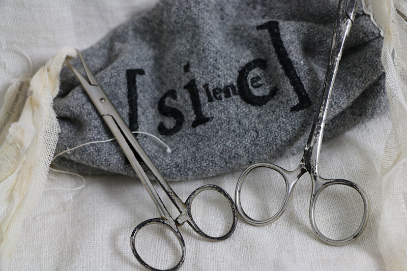 apology artwork .. a full image of the work .. a thin wooden vase of paper flowers sits to the right of frame on a black surface in front of a white textured wall ... the wooden vase has circular markings along its short conical base that straightens into a tall cylinder with notches up its spine .. this vase was once a wooden wool bobbin .. a tag, thin and rectangular, has been affixed near it’s base .. on it, some indistinguishable text ... the tag, with a hole punched in one end, is tied to the vase by twine ... at the top of the vase, long thin wire stems fan out holding a bunch of crisp folded paper flowers formed from book pages. From right to left their faces point out toward the viewer . point up . tilt to one side or another . face the viewer . until the three on the very left tilt face down on slightly curved stems ... some flowers have round blue fabric buttons at their centre .. some have a hint of red thread instead ... on the wall, between the flowers and the base of the vase, are sprinkled shadows .. these shadows from the flowers form a loose arrow shape near the base of the work .. towards clearer star-like shapes at the top