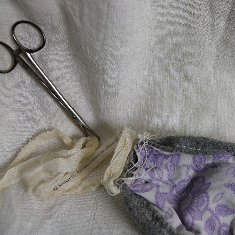 blanket silence artwork detail .. a close up image of the work resting on cream linen fabric ... a pair of silver surgical scissors point down from the top left .. they are gripping two age-stained gauze ties .. on one of the ties some words are visible, printed in black, it reads: 42. Secrecy (1) A person commits an offence if ... the ties are attached to a mask which is laying face down, edges of grey wool rolling over from the front show hand stitching ... the interior of the mask is cotton, a print of purple paisley on white background, fabric which was previously the artist’s pillowcase