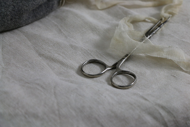 blanket silence artwork detail .. one pair of silver surgical scissors rests on linen fabric ... an edge of the grey woollen face mask is barely visible in the top left corner... the scissors are most prominent in the centre of the image, their pointy end pointing up into the top right, a trail of cream gauze ties in their grip .. the tie flows across from the grey mask on the left .. to the tip of the scissors .. around the left of them then down over their tarnished silver body and off out of sight to the right