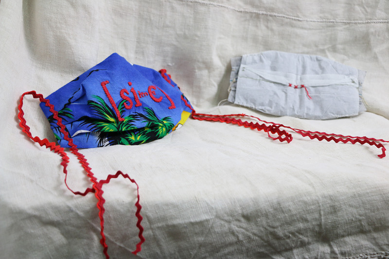burning silence..  a full image of the work .. a hand stitched face mask and filter insert are displayed on a piece of cream linen ...  on the right of the image the white filter sits further back and out of focus .. it has a paper-like cover .. at its centre are 3 red cross stitches and a loose draping thread ... to the front of frame on the left is a brightly coloured face mask resting on an angle ... the body of the mask is formed from reclaimed fabric: blue sky . green palm trees . spots of yellow . white . flying bird silhouettes in the sky .. fabric reclaimed from a Hawaiian shirt ... text has been hand embroidered to its front, vibrant red letters .. [sic] .. with some smaller, indistinguishable letters added in between ... lengths of wavy red ric rac trim are sewn to the mask’s sides to form ties, they drape down from both sides of the mask curving and looping organically as they go