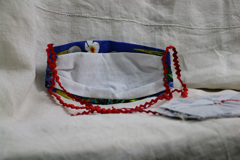 burning silence .. this image focuses on the interior of the mask .. sitting upright on a piece of cream linen the mask is facing away, showing its white inner lining ... red ties hang down from each side to pool together neatly in front ... the filter lays out of focus to the right ... the mask’s body, made from reclaimed Hawaiian shirt, frames the lining with bright blue .. a white flower peeks out from the blue border above the stark white cotton lining .. bold yellows and green feed in from below ... the white lining, attached at the sides and base of the mask, is crisp white cotton .. was once an industrial offcut from a bedding factory .. this lining looks plain . blank . at first .. but closer inspection reveals indentations . with a feint blue.ish tone . what they are is uncertain
