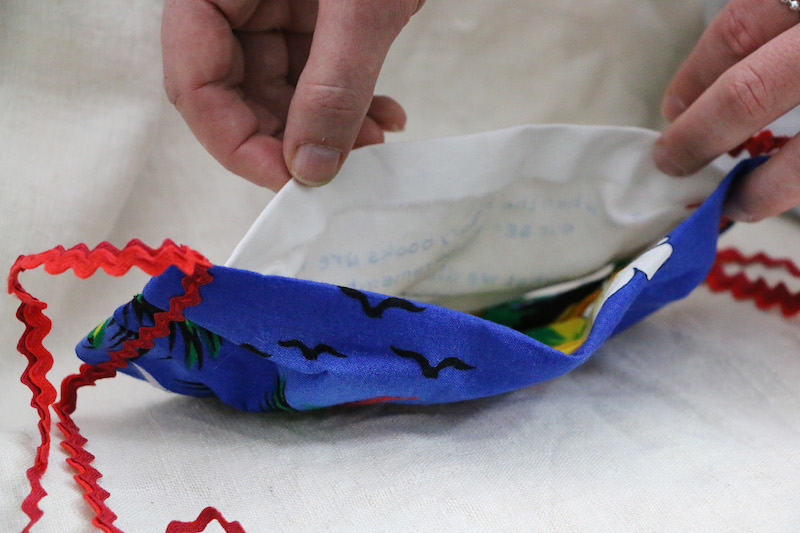 burning silence artwork detail .. the blue face mask lays face down on a piece of cream linen .. the artist holds the back of the mask open .. from this angle, the printed silhouette of 3 birds can be seen on its front .. wavy red ric rac ties flow out to the sides ... the artist's hands reach down from the top .. delicately holding the mask's lining open to reveal the hidden inside ... pale carbon-copy blue markings are visible on the white cotton . the text is unintelligible .. on this back side the text appears like a mirror image .. written in reverse