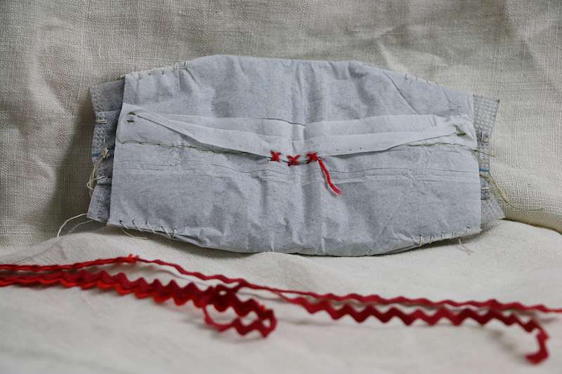 burning silence .. feature image of the charcoal filter on a piece of cream linen . the mask’s wavy red ric rac ties flow across the base of the image from left to right .. slowly blurring as they go ... the filter, sits upright in full focus .. it has a tailor made shape . rectangular . but clipped and curved at the edges . made to fit neatly within the mask .. its main body is formed from non-woven polypropylene fabric reclaimed from a wound dressing .. its sides are crimped and melted shut .. white and cream threads drape from the stitches in its sides ... its body . though white . is darkened by its filling . charcoal . black cinders from a property lost in the black summer bushfires .. the filter's main body is dressed . covered by a thin delicate protective paper sleeve .. its corners each shaped by a run of 5 cream stitches .. this paper joins and is stitched closed front and centre .. one end folds open and is held that way by three red wool symbolic stitches and a draping thread to honour the 33 people directly lost to the fires: x x x...