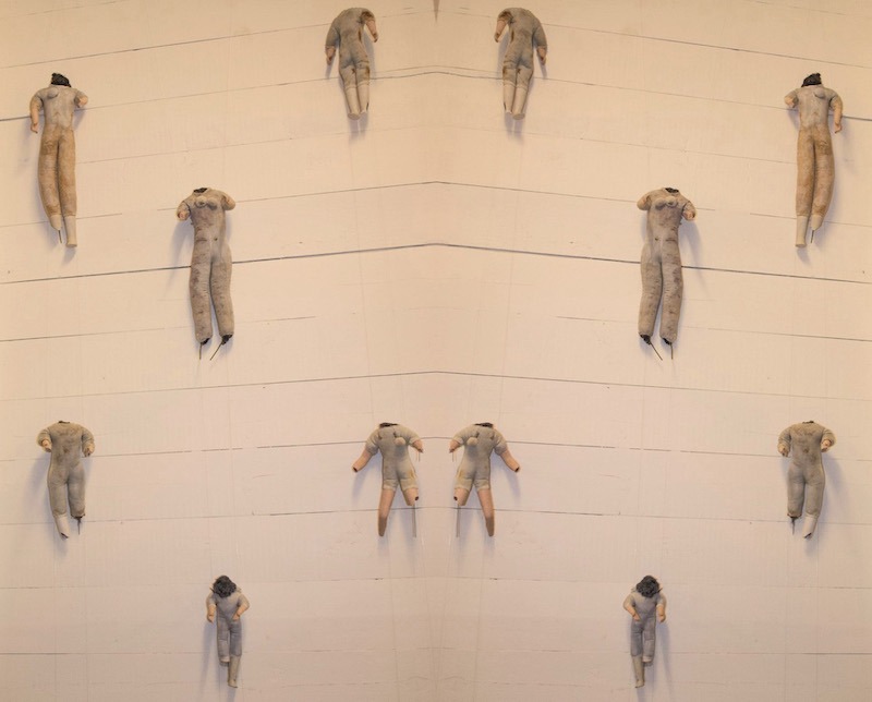 Child Protection. Act. artwork detail .. a mirror image of the dolls against a pale wooden wall of horizontal slats .. headless vintage dolls are hanging almost equally spaced . connected by barely visible threads .. the photo has a pinkish golden tone ... six headless dolls are reflected by the mirroring so we see twelve ... they all have fabric bodies and ceramic pale limbs . their bodies are stained and worn . some have dark stuffing coming out of the neck .. none have heads ... many limbs are broken . their arms reach out towards the viewer ... the top two . shown on a slight angle . with arms and legs straight down .. look like they are flying upwards