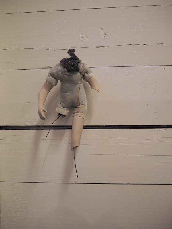 Child Protection. Act. detail of one headless doll on a wooden slatted wall .. the photo has a warm pinkish golden tone .. this doll looks full of stories ... its pale fabric body has a straight rust-coloured stain across the left torso to shoulder .. this doll has an interesting almost active . almost running . shape ... its left leg is broken off at the thigh leaving a short piece of wire almost searching sideways .. the right is ceramic until broken off and becoming wire at the knee .. its right arm is ceramic from the upper arm until broken off at the elbow .. its left fully intact . with palm facing the body . is hanging down in an active curve ... lots of dark stuffing is coming up from the neck . one piece is curled up almost like a baby swan's neck ... this doll is full of stories
