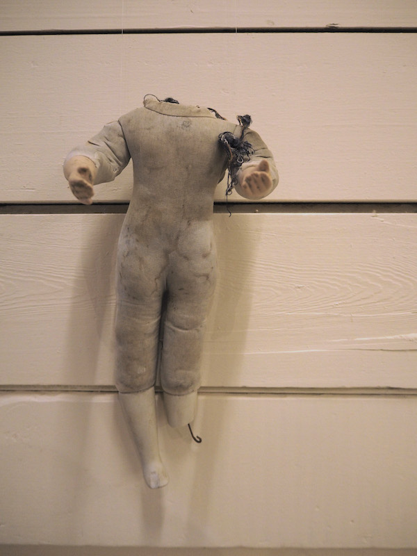 Child Protection. Act. detail of one headless doll on a wooden slatted wall .. the photo has a warm pinkish golden tone .. this doll looks full of stories ... its greyish fabric body is stained .. by muck and moisture .. ceramic legs are at an angle . one broken with a piece of hooked wire hanging from the break .. both arms are reaching forward .. left palm facing up .. an accumulation of threads hangs from its shoulder ... this doll is full of stories
