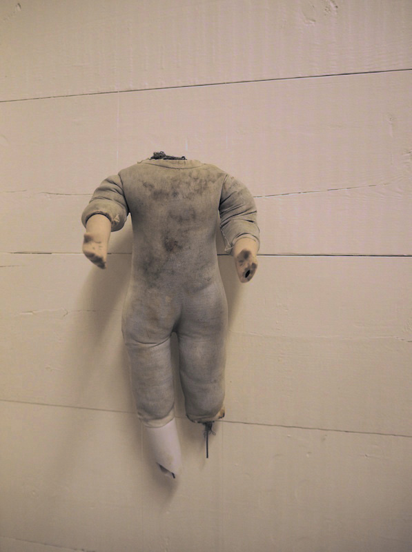 Child Protection. Act. detail of one headless doll on a wooden slatted wall .. the photo has a warm pinkish golden tone .. this doll looks full of stories ... its greyish fabric body is stained .. by muck and moisture .. ceramic legs starting at the knee are broken . one is missing with a short piece of wire pointing down from the break .. the other is broken at the ankle .. both arms are reaching forward .. fingers on the left hand have broken off .. the right palm faces up ... this doll is full of stories