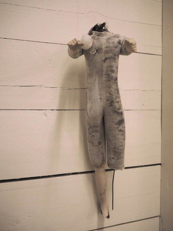 Child Protection. Act. detail of one headless doll on a wooden slatted wall .. the photo has a warm pinkish golden tone .. this doll looks full of stories ... its greyish fabric body is stained right down its right side and left thigh by dark muck .. one leg is broken off at the knee with only wire in its place .. the other is broken off at the foot .. one padded circle is attached right side of the chest .. some dark stuffing is coming up from the neck ... both arms are reaching forward .. palms tilted toward each other and down ... this doll is full of stories