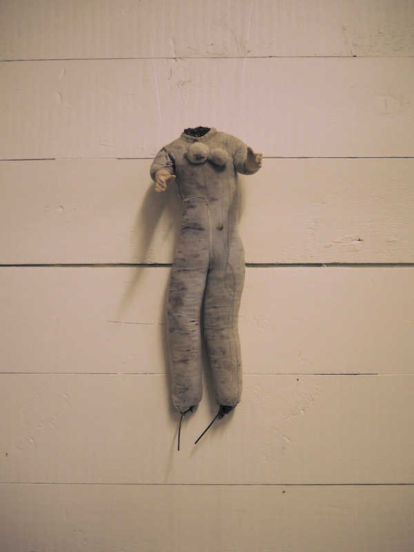 Child Protection. Act. detail of one headless doll on a wooden slatted wall .. the photo has a warm pinkish golden tone .. this doll looks full of stories ... its greyish fabric body is stained .. dark muck in places .. its legs are wide and long .. fabric stuffed and equally shaped to the ankles where stuffing and angled wire replace feet .. high on the chest are two padded circles . their location a bit out of place ... both arms are reaching forward .. one slightly above the other as if for a hug ... this doll is full of stories