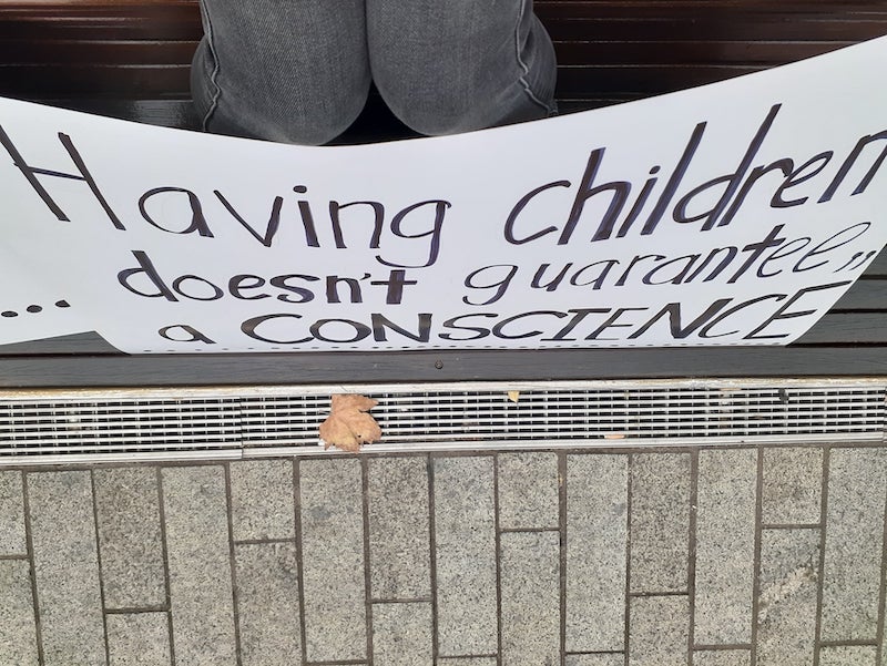an image of the poet and artist indigo eli sitting on a dark brown slatted bench . with a placard of Grace Tame’s words resting against her knees . she is wearing black denim jeans .. the image has been taken by the artist from above ... thin grey vertical pavers line the bottom of the image .. above them a horizontal silver drain is adorned with one fallen autumn-orange oak leaf .. above that is a large white card covered with black handwritten words. It reads: ... Having children doesn't guarantee a conscience"