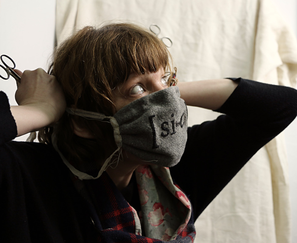 a playful headshot of the artist and poet indigo eli .. a Caucasian woman with golden brown hair cut with a fringe ... she is wearing a black jumper pulled up at the forearms and a floral & tartan scarf .. she is holding a textile artwork, a wearable document, around her face .. a grey woollen mask that has [sic] embroidered in black letters on the front .. it covers her nose and mouth .. it’s gauze ties pull back to each side where a pair of scissors are held loosely by their points in each fist .. the round handles stand to attention like ears ... her body is turned slightly .. her eyes are cast to the side ... and up