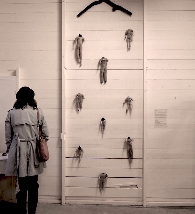 Child Protection. Act. artwork .. a full image of the work . a wall hanging on display ... a white wooden wall . built from horizontal slats . is divided by two thin vertical studs seperating the wall . and this image . into three vertical sections .. the photo has a warm pinkish tone ... a woman stands on the left facing the wall . or the work . it is difficult to determine exactly where her gaze rests .. she is wearing black pants . a pale blue structured canvas jacket with ties hanging from the back .. a tan handbag over her right shoulder .. shoulder length dark hair and a black hat .. some other work is slightly visible on a curved white table to her left ... the right section is blank apart from an artwork description from this distance it is unreadable ... in the middle section the art work fills the wall . at 3 metres it hangs at almost twice the height of the lady .. this work is made from machinery and vintage dolls .. at the top . a flat dark piece of metal . an industrial looming jack from a woollen mill . looks a bit like a coat-hanger .. under this metal frame hang nine headless dolls equally spaced .. from the top one hands from each end .. then below them . one hangs in the middle .. then two .. then a little one .. two again .. and then one