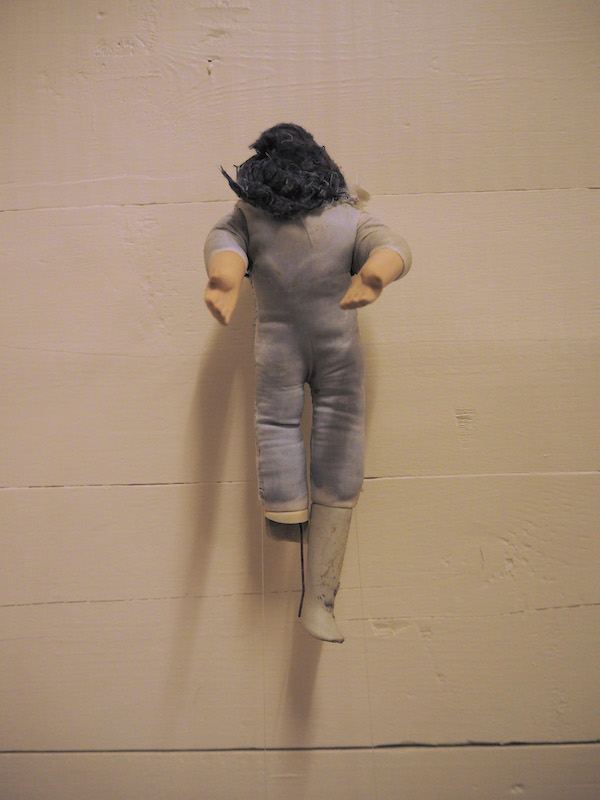 Child Protection. Act. detail of one headless doll on a wooden slatted wall .. the photo has a warm pinkish golden almost pearlescent tone .. this doll looks full of stories ... its pale fabric body shows the dark stuffing underneath ... its left leg . white ceramic boot from the knee . is fully intact with some dark stains .. the right is broken off at the knee with a dark wire hanging straight down in line with the left leg .. its arms, pale ceramic from the elbows, are reaching forward toward the viewer . the doll’s left palm is facing up ... dark stuffing . rising up from its neck into a roundish heap with some wisps off to the side . is shaped a bit like soft serve ice-cream ... this doll is full of stories